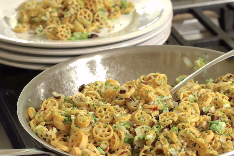 Wagon Wheel Chickpea Pasta tossed with sautéed veggies in a skillet; serving on plate in the background