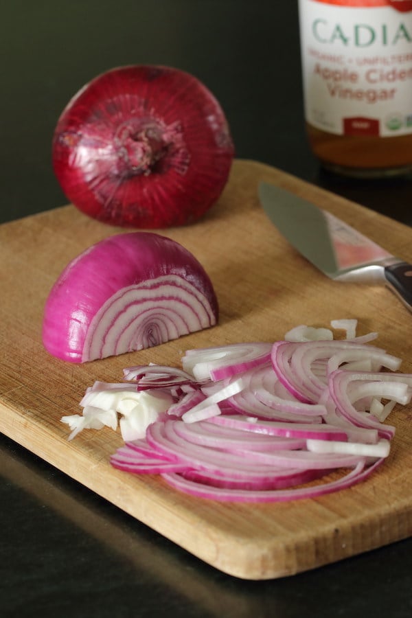 A whole red onion, peeled half red onion, and sliced onion on a cutting board; a bottle of apple cider vinegar is in the distance