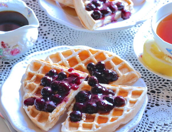 Plates of crispy gluten-free vegan waffles with blueberry sauce, herbal tea, and pitcher of maple syrup