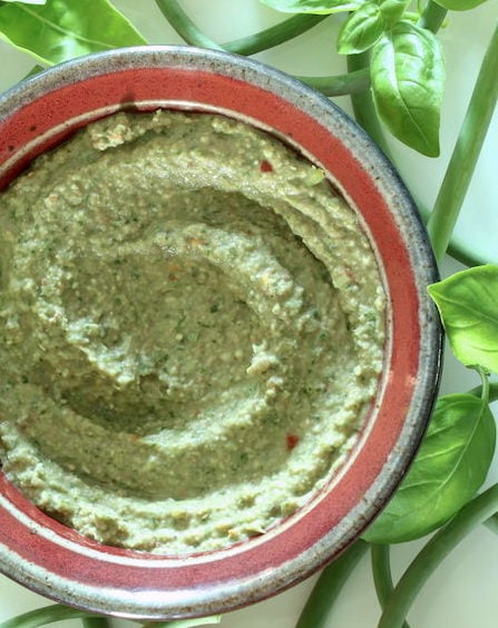 Oil-free vegan pesto in a small bowl against a white background with basil leaves and garlic scapes strewn around