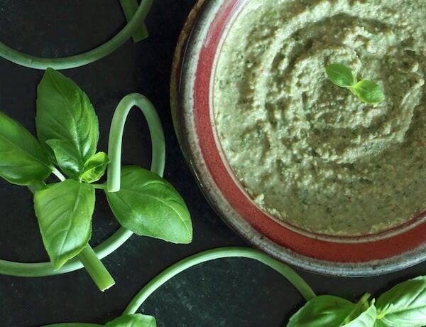Bowl of Oil-Free Vegan Pesto surrounded by garlic scapes and basil leaves
