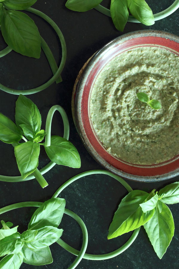 Bowl of Oil-Free Vegan Pesto surrounded by garlic scapes and basil leaves