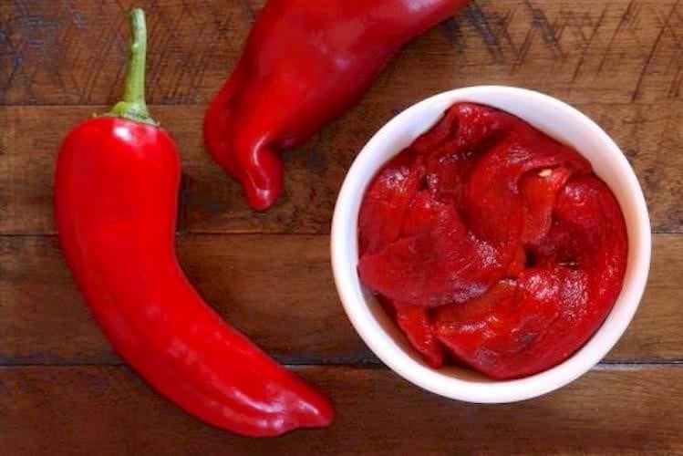 Two fresh red peppers arranged around a small bowl of roasted red peppers