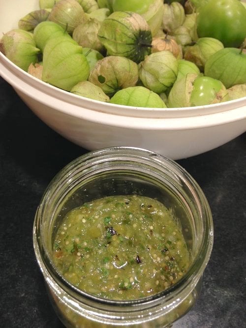 In background, a bowl of tomatillos with husks still intact. In foreground, a jar of tomatillo salsa