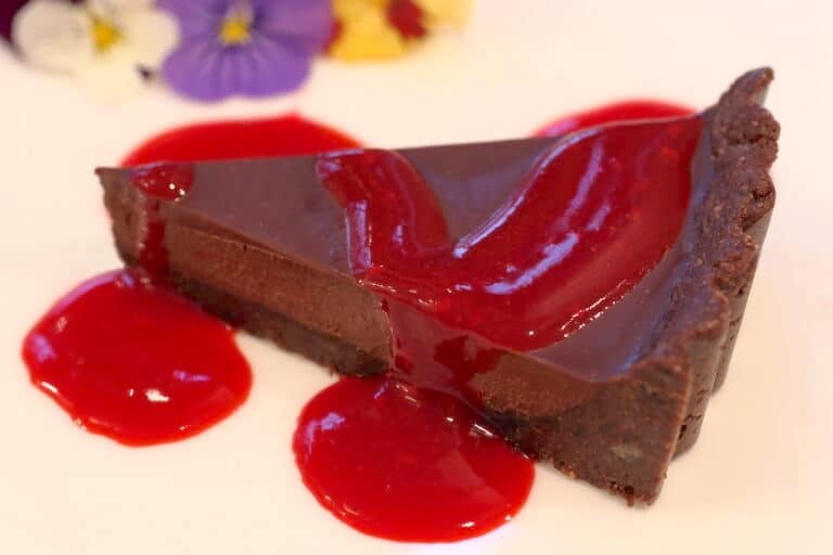 Close up of a slice of Vegan Chocolate Tart with raspberry sauce; edible pansies in the distance